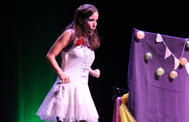 Mujer sobre un escenario vestida con un disfraz blanco tipo hada o personaje de cuento, con maquillaje llamativo y un micrófono de diadema. A su lado, hay una estructura cubierta con una tela morada decorada con bolas y guirnaldas de banderines