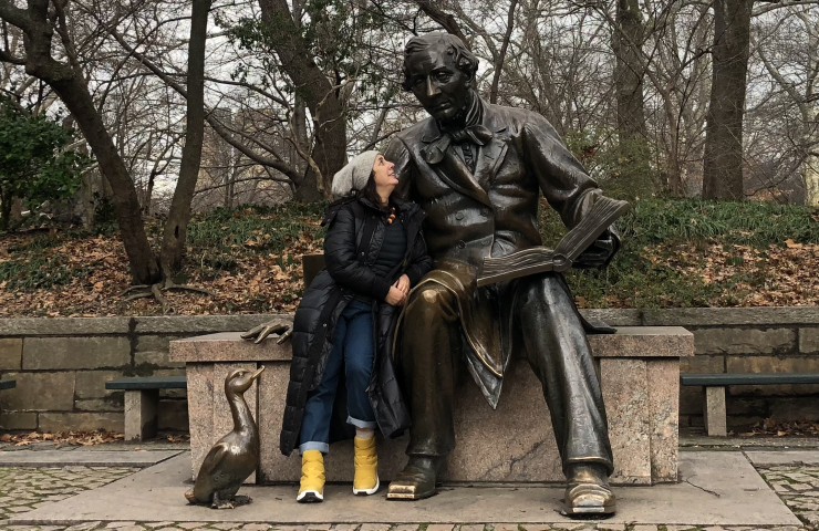 Mujer vestida con abrigo y gorro de invierno, posando junto a una gran estatua de bronce de un hombre sentado con un libro abierto y una figura de pato cerca. La estatua representa al escritor Hans Christian Andersen.